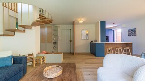 A modern living room with a wooden staircase, a sofa, a coffee table, and a kitchen area with stools. - Sea-View Duplex for Family (Villeneuve-Loubet)