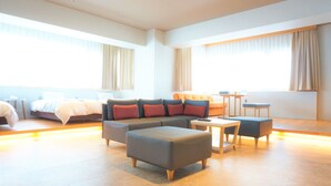 A modern hotel room with a large bed, a grey sofa, a small table, and a seating area with ottomans. - Yukureba Maejima Hotel (Naha)