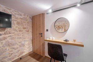 A compact room with a stone wall, a wooden door, a round mirror, a small desk with a chair, and a flat-screen TV mounted on the wall. - Pileas Boutique, Apartment (Aegina)