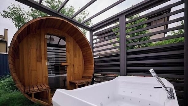 Una bañera de hidromasaje de madera con un banco en el interior de una estructura circular de madera, una bañera de hidromasaje exterior blanca y una barandilla de metal.