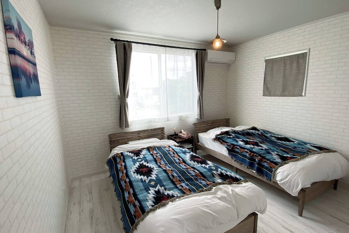 Een kamer met twee bedden, witte bakstenen muren, een raam met gordijnen en een hanglamp.