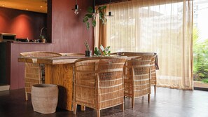 Un coin repas avec une table en bois et des chaises en osier, un petit coin cuisine équipé d’un four à micro-ondes, et une grande fenêtre ornée de rideaux légers.