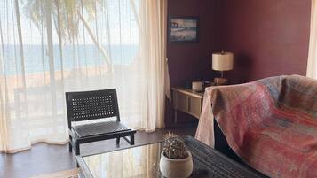A room with a view of the sea, a chair, a table with a potted plant, a lamp, and a bed with a blanket.