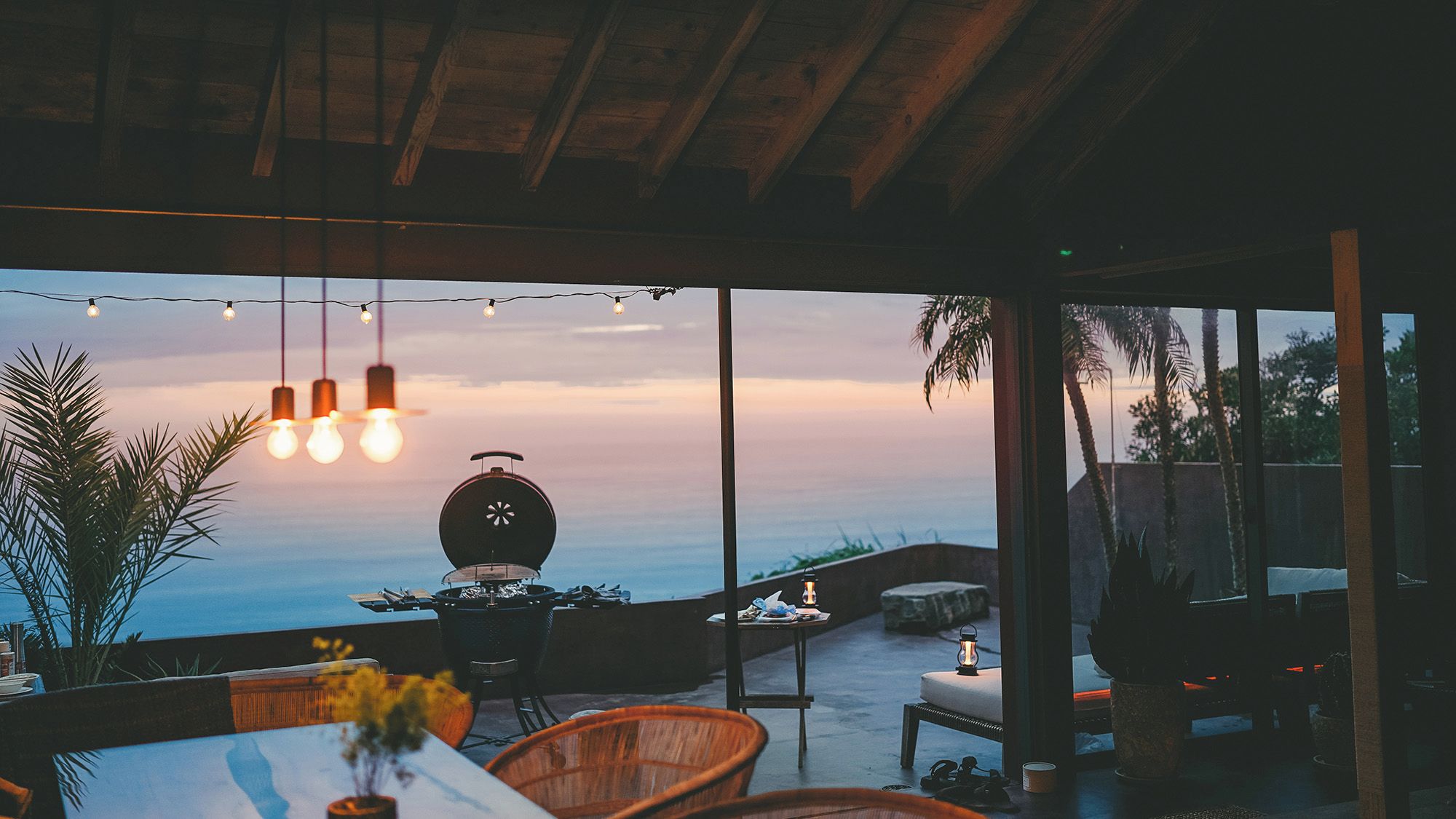 Un área de comedor al aire libre con vista al océano, luces colgantes y una parrilla.