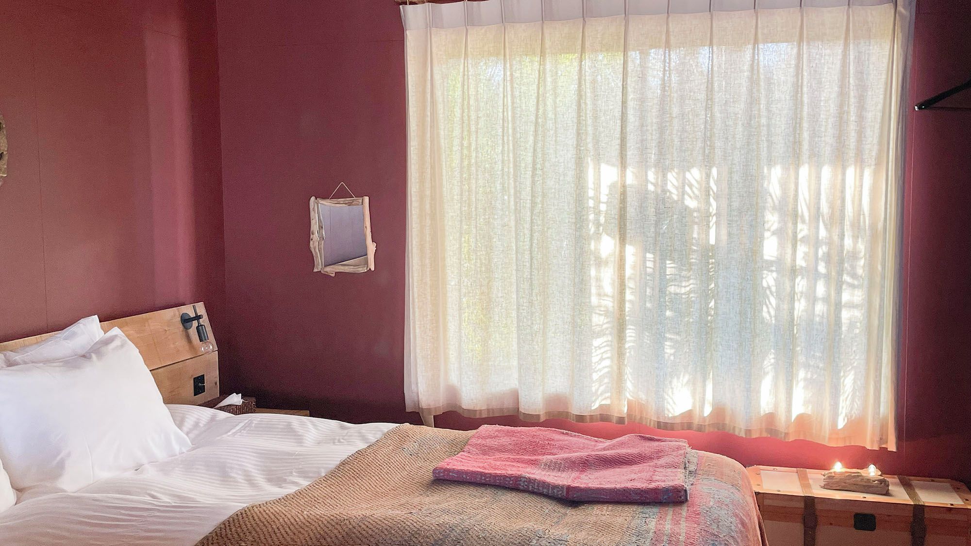 Un dormitorio con cama, una mesita de noche, cortinas translúcidas en la ventana y un espejo fijado a la pared.