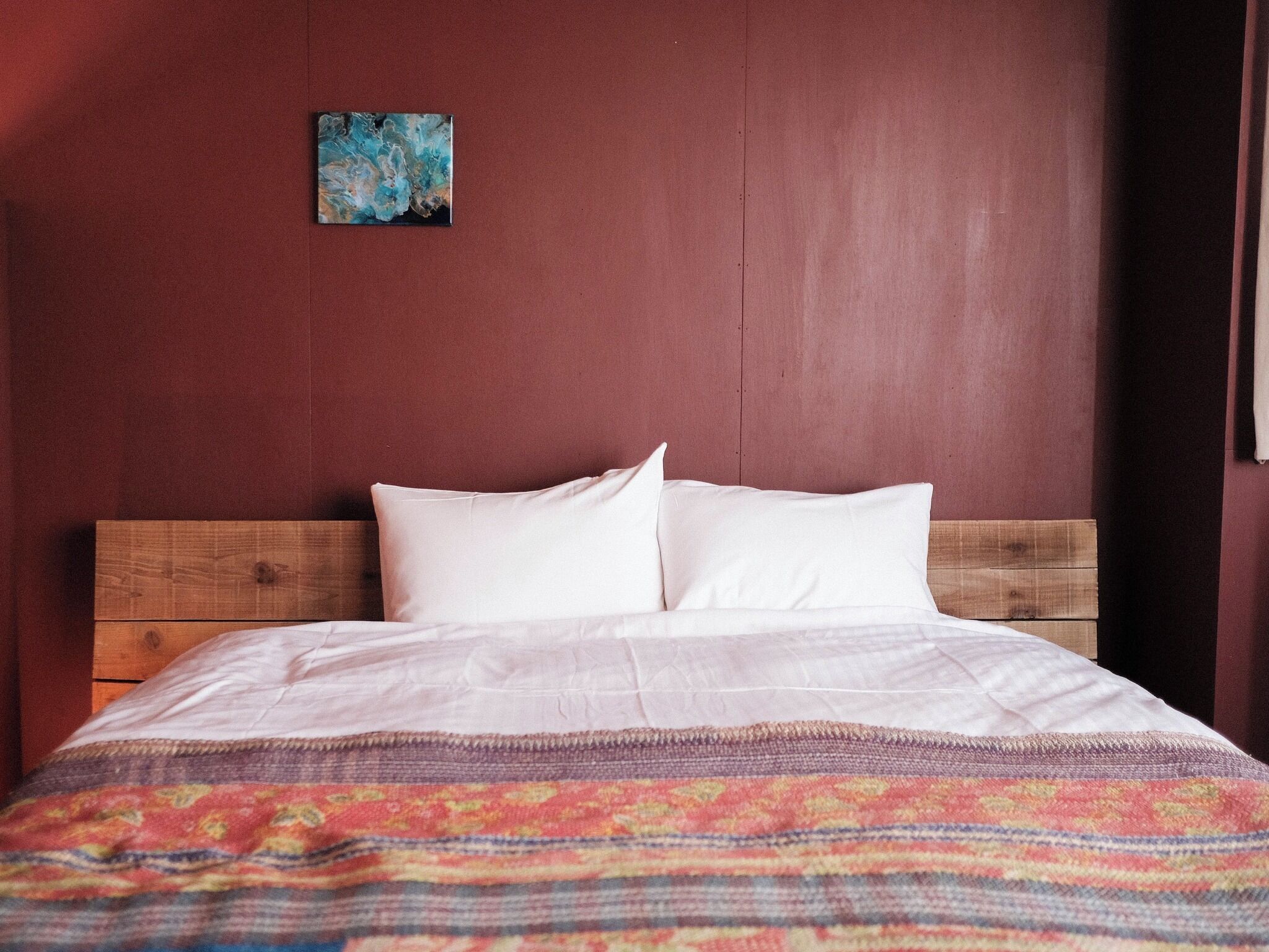 A bed with white bedding and a colorful blanket, a wooden headboard, and a red wall with a small blue painting.