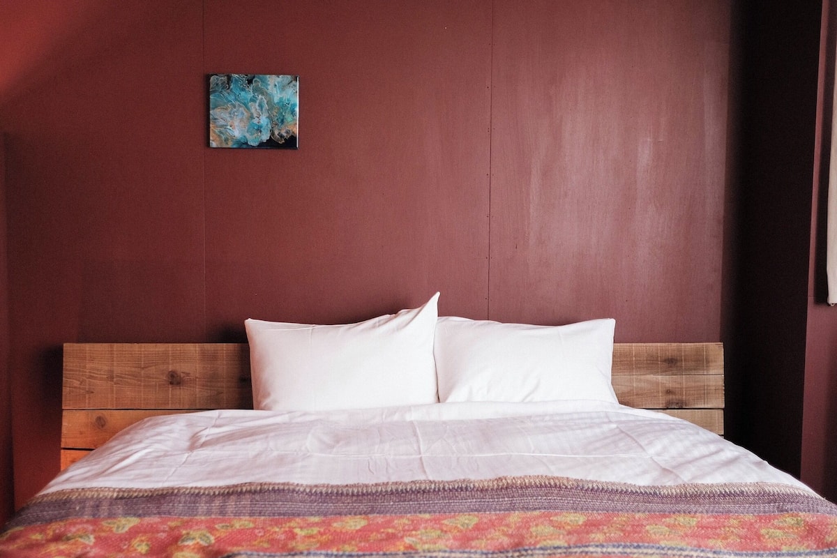 Una cama con ropa de cama blanca y una colcha colorida, un cabecero de madera y una pared roja con un pequeño cuadro azul.