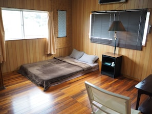 Une petite chambre avec un lit simple, des murs et un plancher en bois, une fenêtre avec des rideaux, une table de chevet avec une lampe et une chaise.