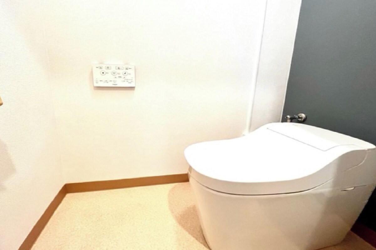 A small, compact bathroom with a toilet, a wall-mounted electrical outlet, and a door with a silver handle.
