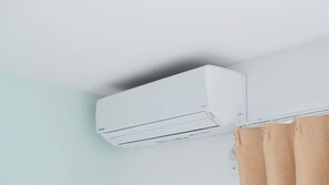 A white air conditioning unit mounted on a wall in a room with light blue walls and a brown curtain. - Condominium Yuyuki (Yomitan)