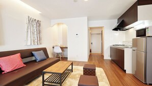 A modern living room with a sofa, coffee table, and ottoman. There is a kitchen area with a refrigerator and microwave. - Condominium Yuyuki (Yomitan)