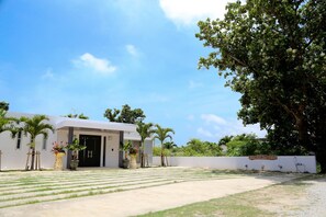 A modern building with a flat roof, a paved entrance area, and a large tree to the right. - Palm Villa Ishigakijima Igusa (Ishigaki)