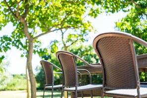 Des chaises et une table en osier, destinées à être placées à l’extérieur, probablement pour prendre un repas ou se détendre, dans un cadre verdoyant qui ressemble à un parc, avec des arbres en arrière-plan.