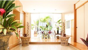 A spacious hallway with a tiled floor, leading to a bright room with large windows and a view of palm trees. There are wicker chairs and potted plants along the hallway. - Palm Villa Ishigakijima Igusa (Ishigaki)