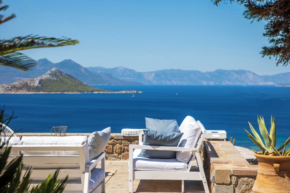 Un salon de bord de mer avec des chaises et des coussins blancs, une plante en pot et une vue sur la mer et les montagnes.