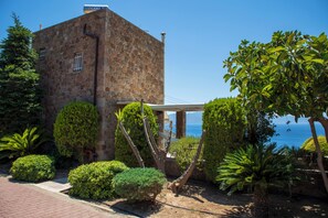 A stone building with a view of the sea, surrounded by well-maintained bushes and a clear sky.