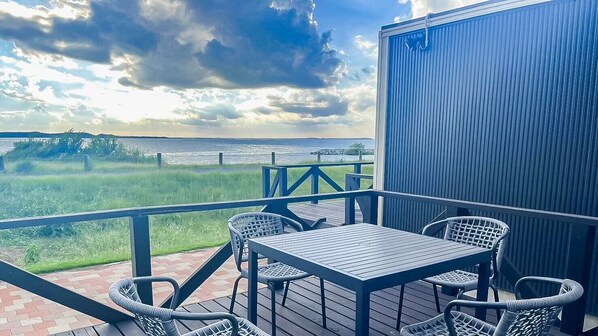 Une terrasse avec une table et des chaises, donnant sur une étendue d’eau et une zone herbeuse.