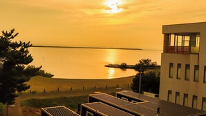 Ein modernes Gebäude mit flachem Dach und großen Fenstern, mit Blick auf ein ruhiges Gewässer bei Sonnenuntergang.