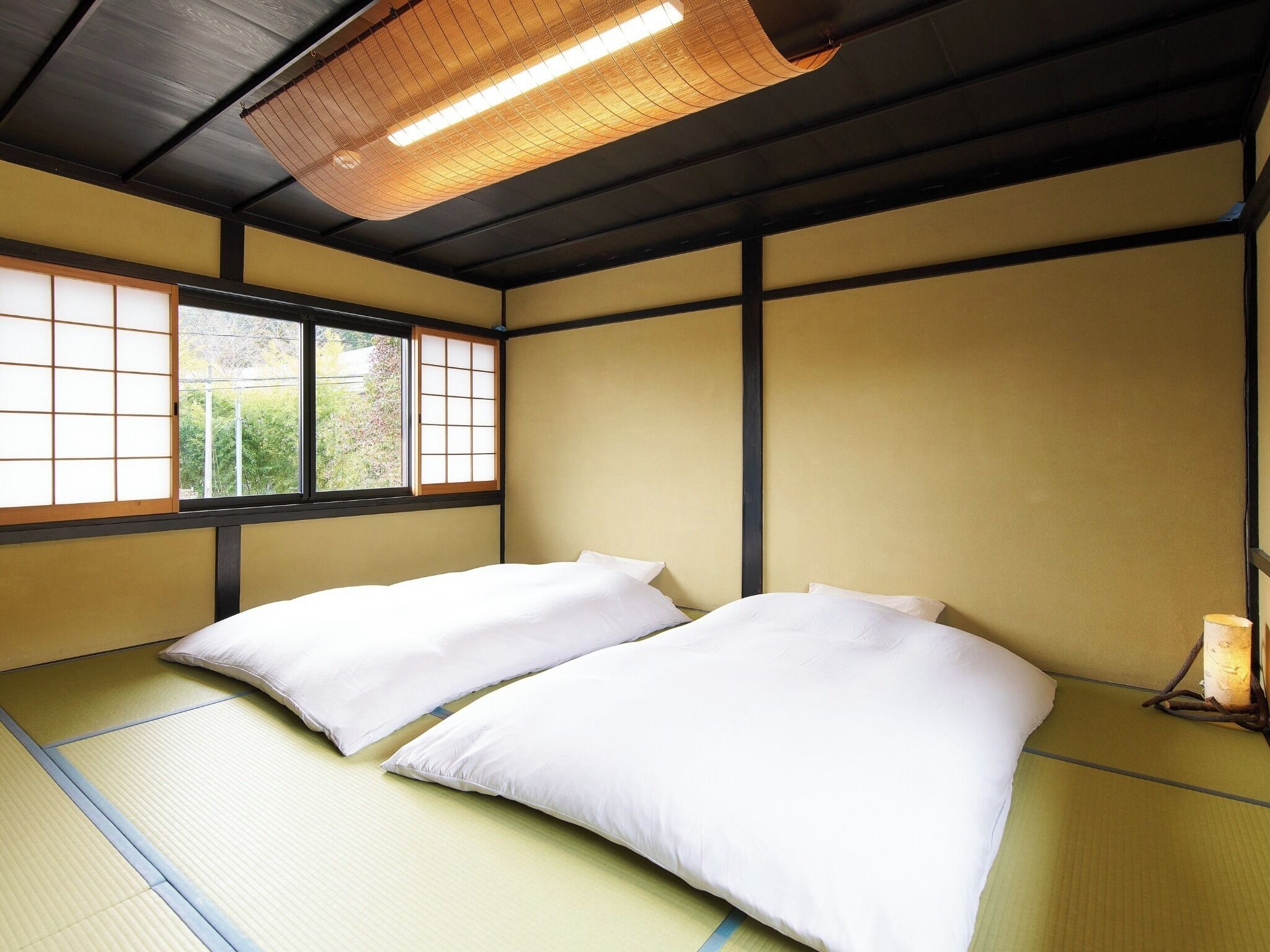 A traditional Japanese-style room with tatami flooring, two white beds, a shoji screen window, and a hanging light fixture.