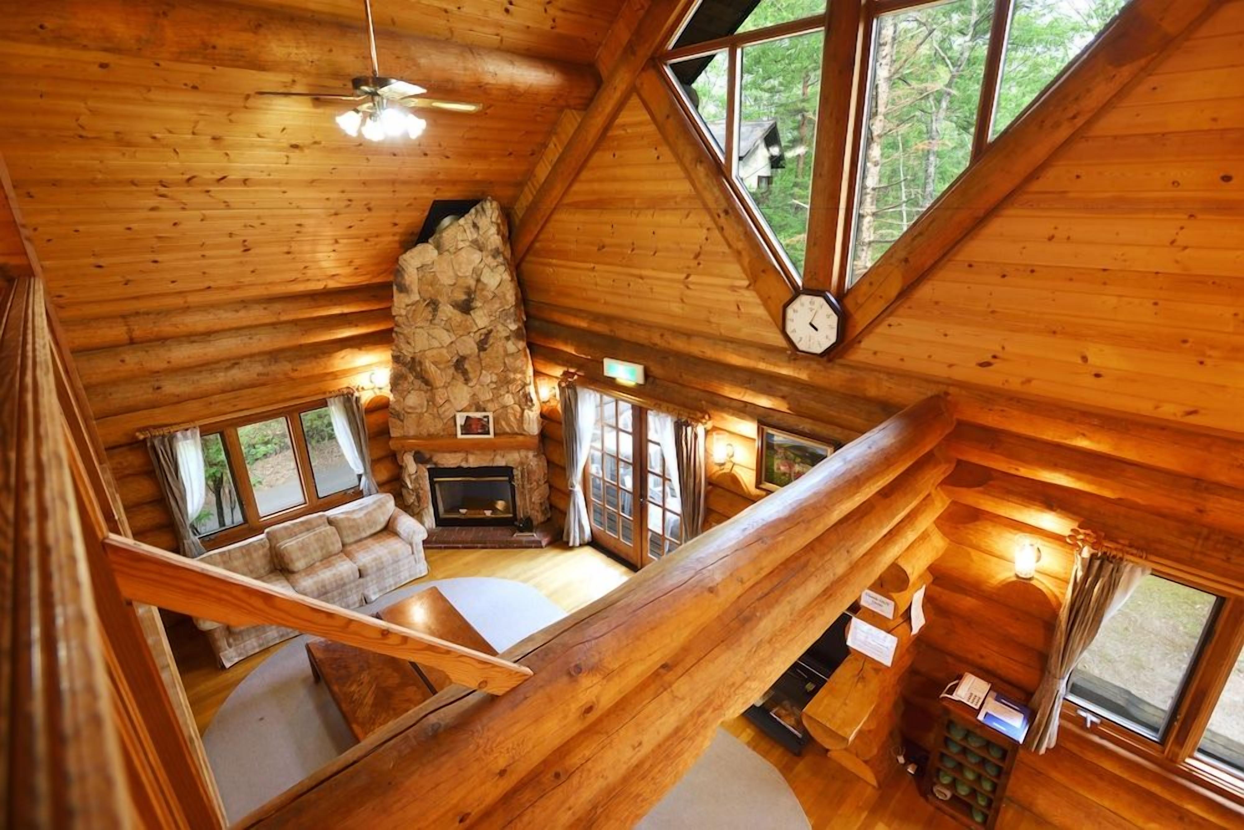 A log cabin interior with a stone fireplace, a loft bedroom with a wooden staircase, and large windows offering a view of the outdoors.