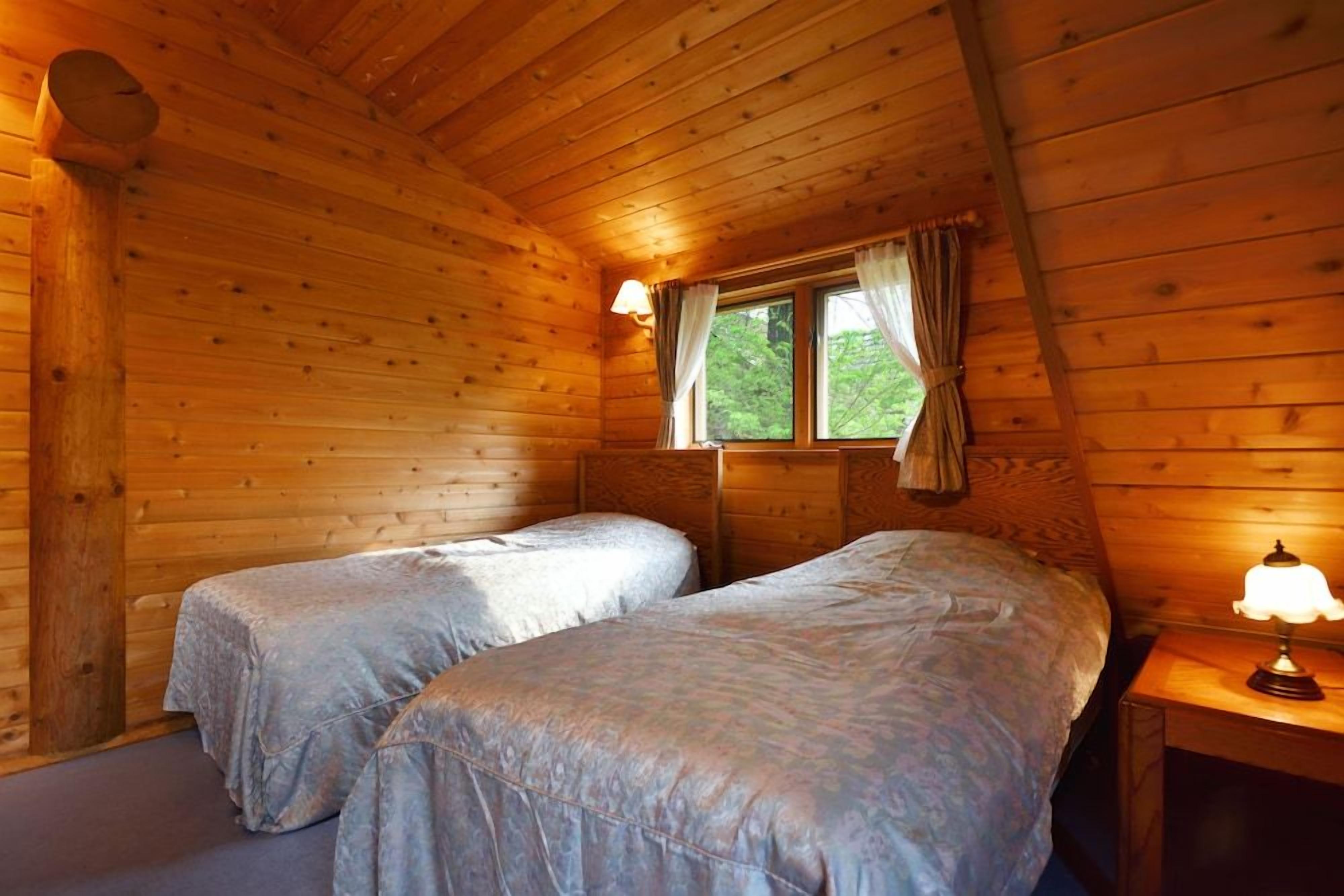 Una cabaña con dos camas, una ventana con cortinas y una lámpara en una mesita de noche.