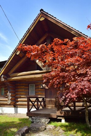 Una cabaña de troncos con un porche de madera y un árbol con hojas rojas al frente.