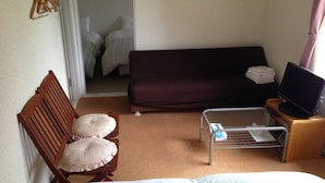 A small, cozy room with a bed, a wooden chair with cushions, a small table with a television, and a sofa.