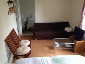 Una habitación con una cama, un sofá, un televisor sobre un mueble, una silla con cojines y una mesita.