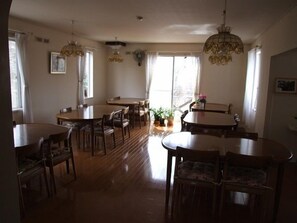 A dining area with multiple tables and chairs, large windows with curtains, and a chandelier.