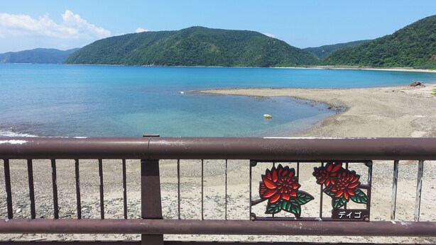 A coastal landscape with a clear sky, a sandy beach, and a mountain in the background.
