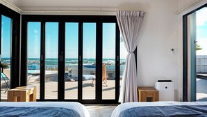 Une chambre d’hôtel dotée d’une grande porte-fenêtre coulissante donnant sur un balcon avec vue sur la plage et l’océan. La chambre comprend deux lits avec du linge de lit bleu, une petite table en bois et un appareil de climatisation blanc.