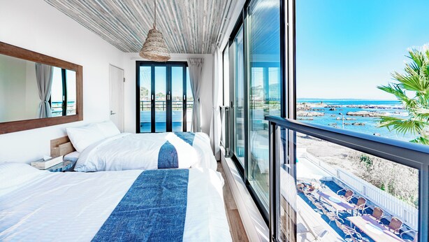 Ein Hotelzimmer mit einem großen Bett, einer Hängelampe und einem Balkon mit Blick auf Strand und Meer.
