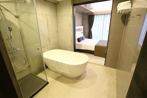 Ein modernes Badezimmer mit einer gläsernen Duschkabine, einer freistehenden Badewanne und einem sichtbaren Schlafzimmer mit Bett und Vorhängen.