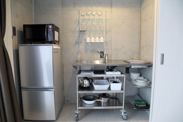Une petite cuisine équipée comprenant un réfrigérateur en acier inoxydable, un four à micro-ondes, un évier et une étagère en verre où sont rangées des tasses et des verres.