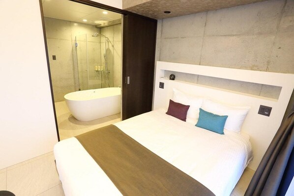 A modern hotel room with a large bed, a separate bathroom with a bathtub, and a glass shower enclosure.