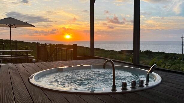 A wooden deck with a hot tub and an umbrella, overlooking a sunset over the ocean.