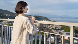 Une personne se tient sur un balcon, d’où elle a une vue sur la ville, avec des bâtiments et des collines qui se dessinent à l’horizon.