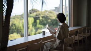 Une personne est assise près d’une fenêtre, un livre à la main, et la vue donne sur des arbres et un paysage urbain.
