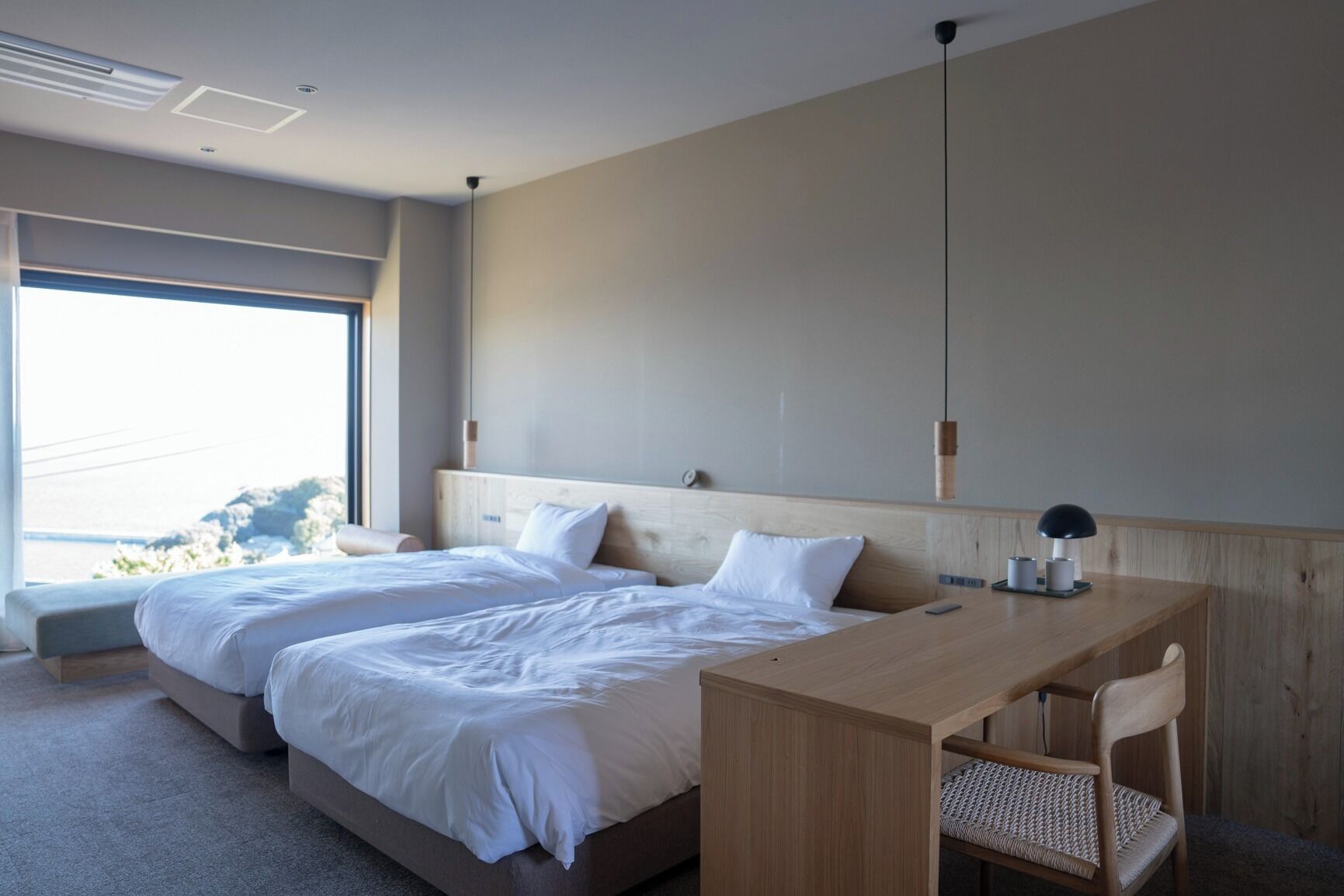 A modern hotel room with two beds, a wooden desk, and a large window offering a view of the outdoors.