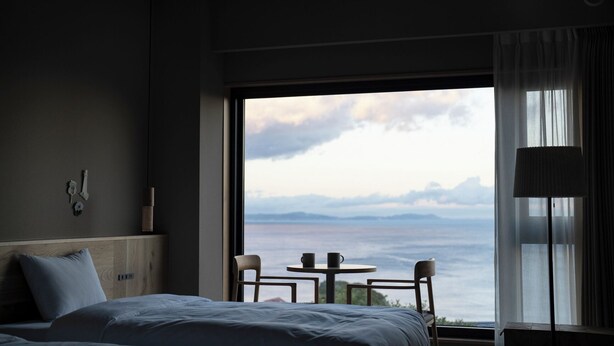 Une chambre d’hôtel avec une grande fenêtre, un lit, une table avec deux chaises et une lampe.