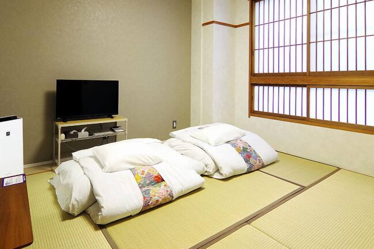 Une pièce de style japonais traditionnel, avec un sol recouvert de tatamis, un téléviseur posé sur un meuble et une fenêtre ornée de barreaux en bois.