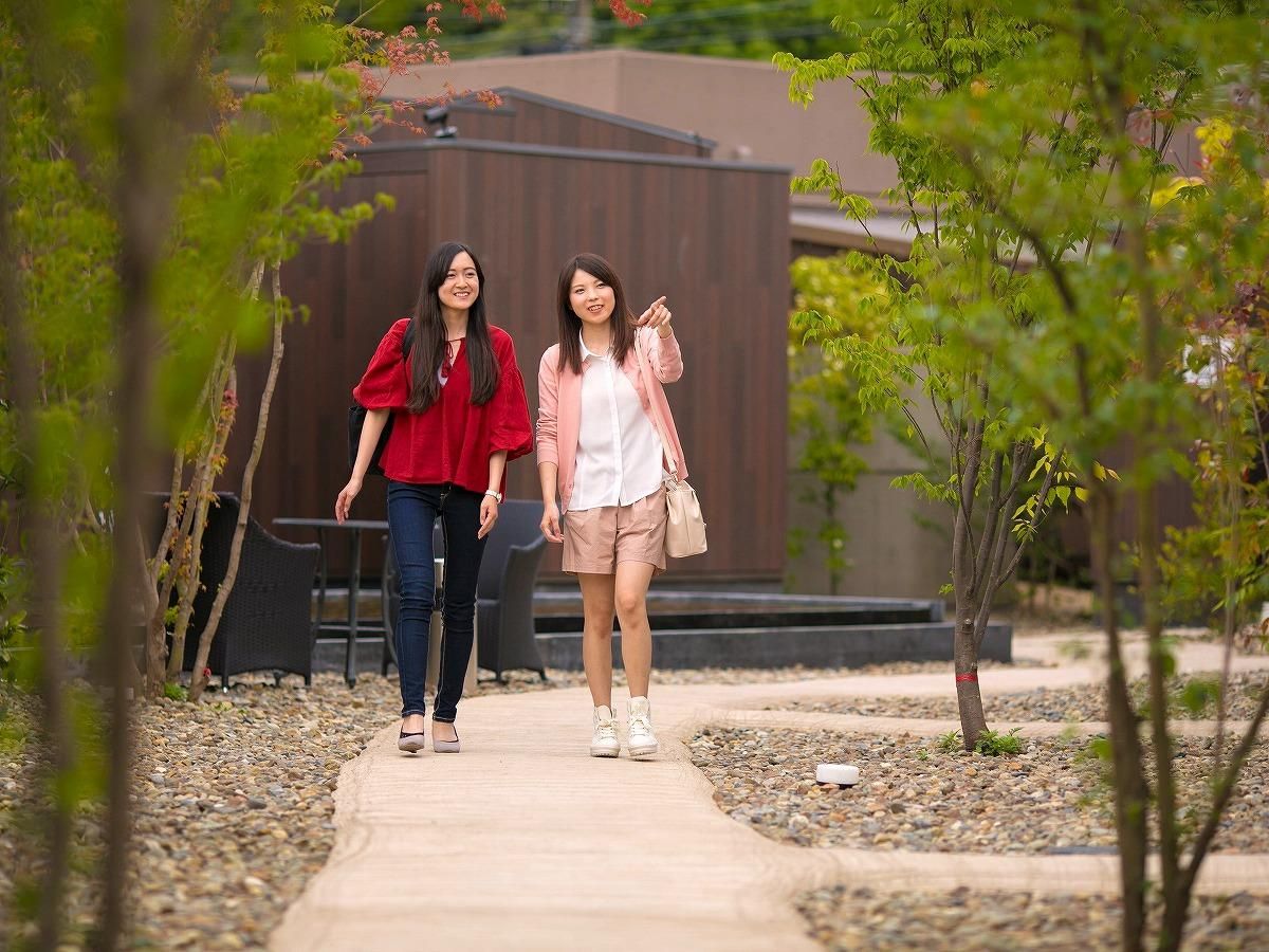 Two individuals walking on a pathway surrounded by trees and a building in the background.