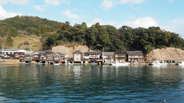 A waterfront area with traditional wooden buildings, boats, and a hillside with dense greenery. - Kominkano Yado Saika-an (Maizuru)