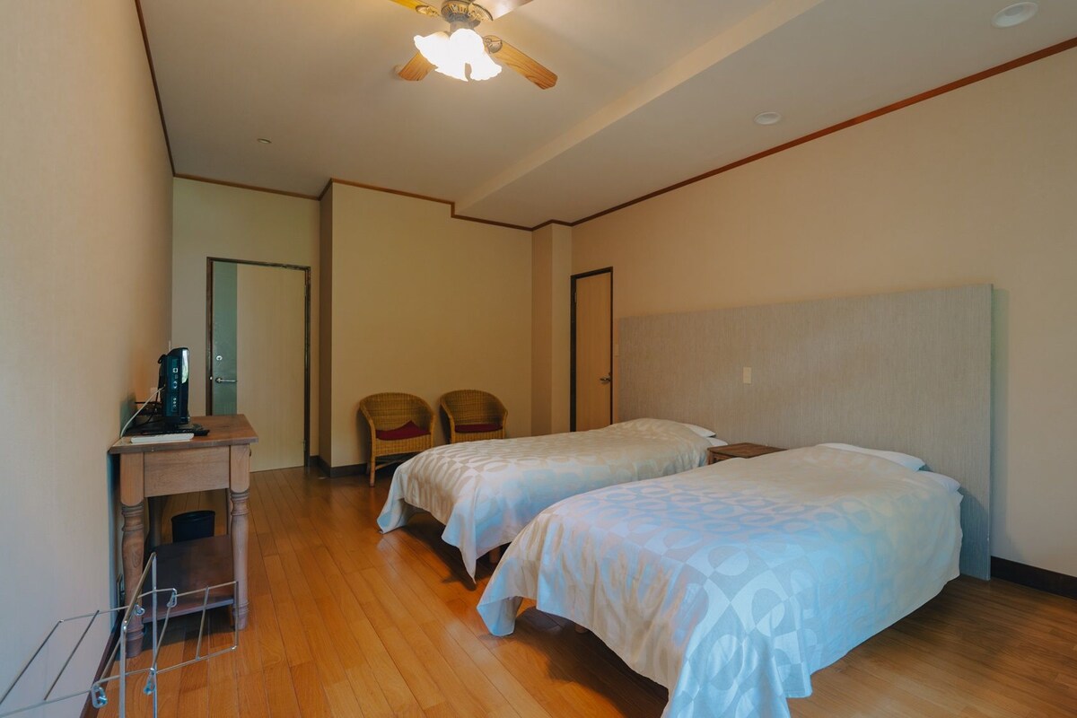 A hotel room with two beds, a desk with a coffee maker, a chair, and a ceiling fan.