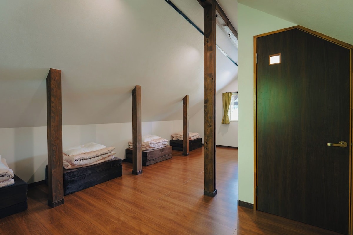 A room with four beds, wooden beams, a wooden door, and a window with curtains.