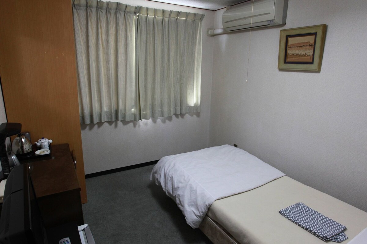 A small, clean room with a single bed, a desk with a kettle and a phone, a window with curtains, and a framed picture on the wall.