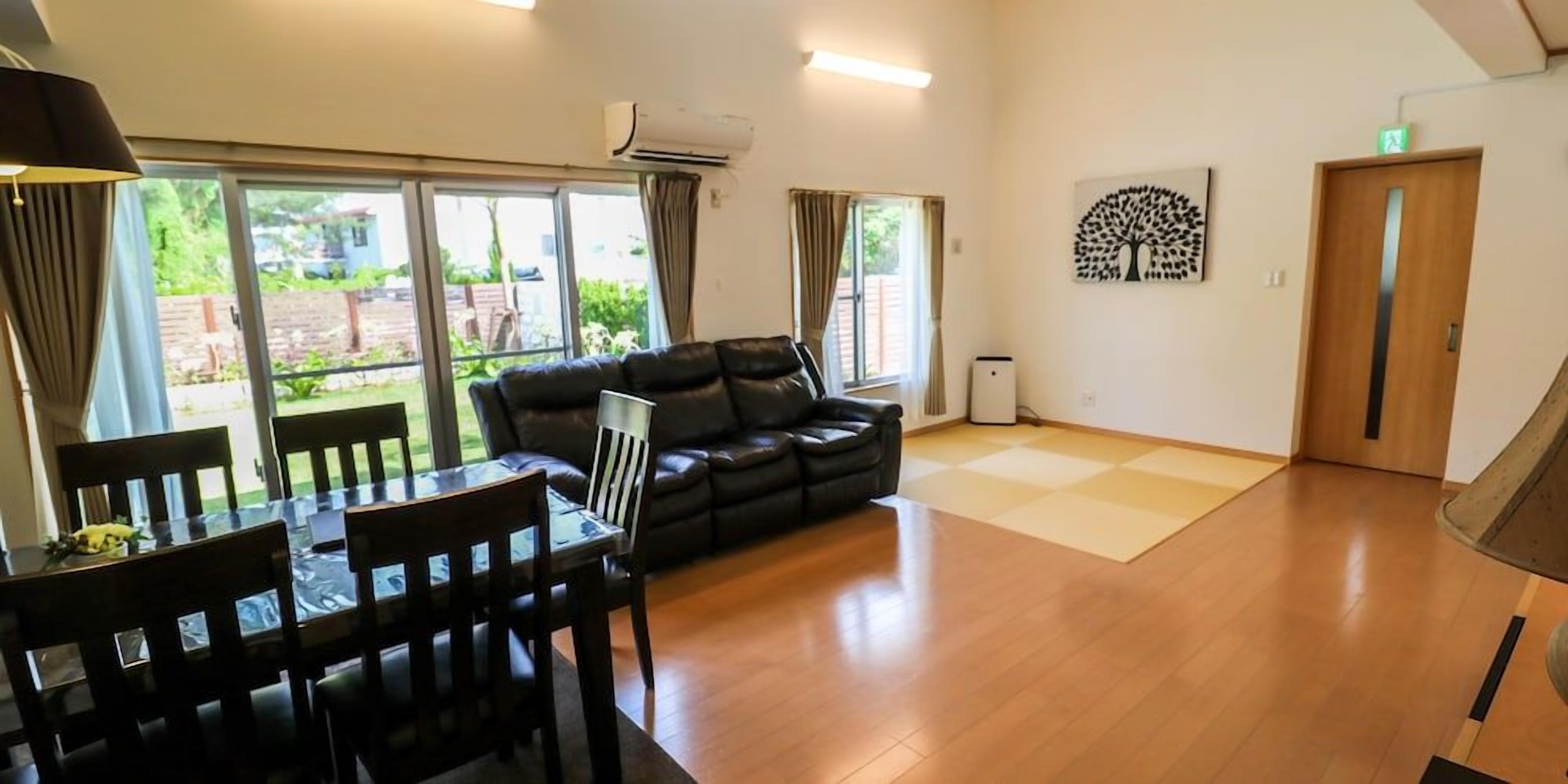 A room with a dining table, chairs, a sofa, and a large window with curtains.