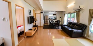 A hotel room with a wooden floor, a sofa, a TV, a dining area, and an air conditioning unit.