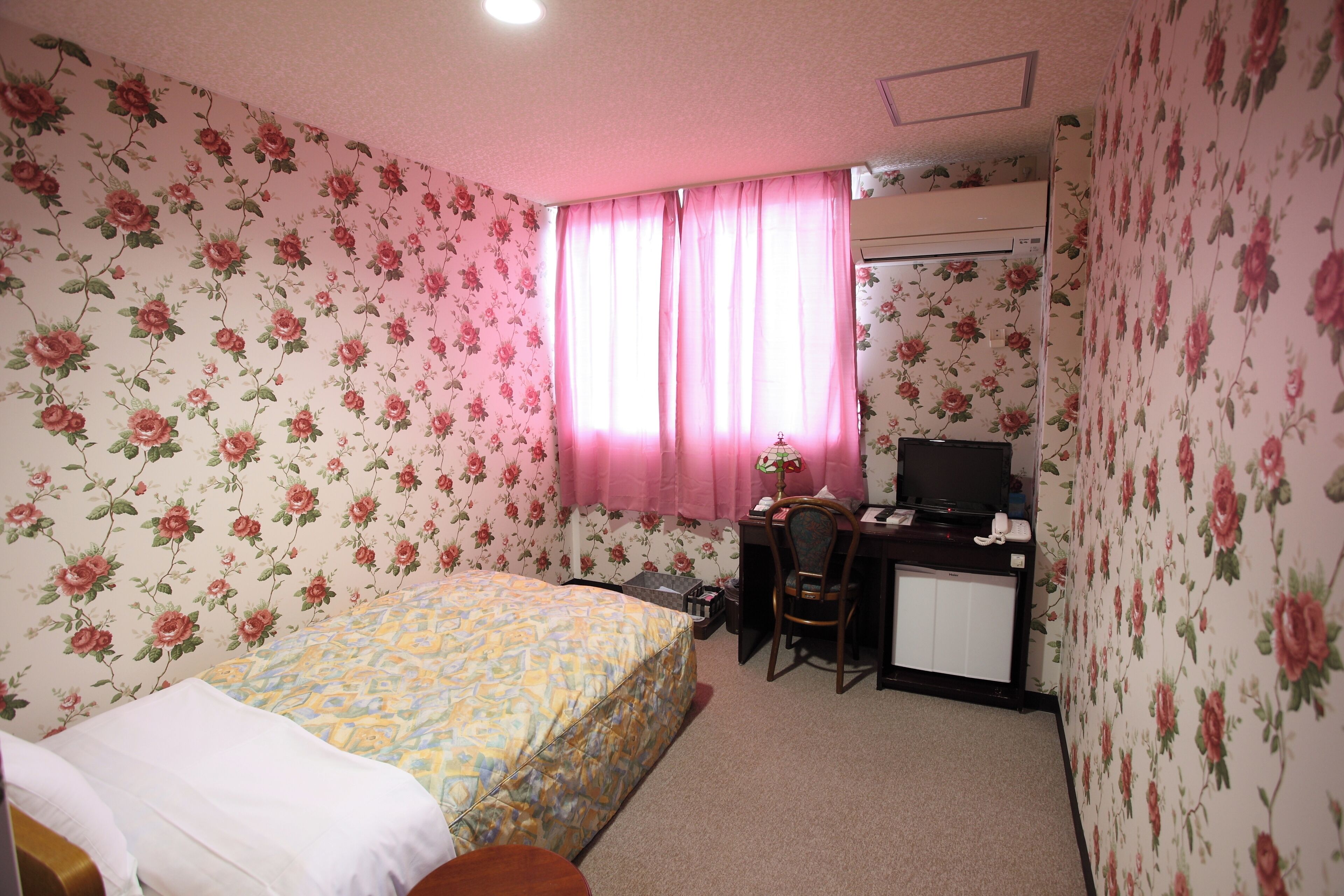 A bedroom with floral wallpaper, a bed with a patterned bedspread, a desk with a chair, and a television.