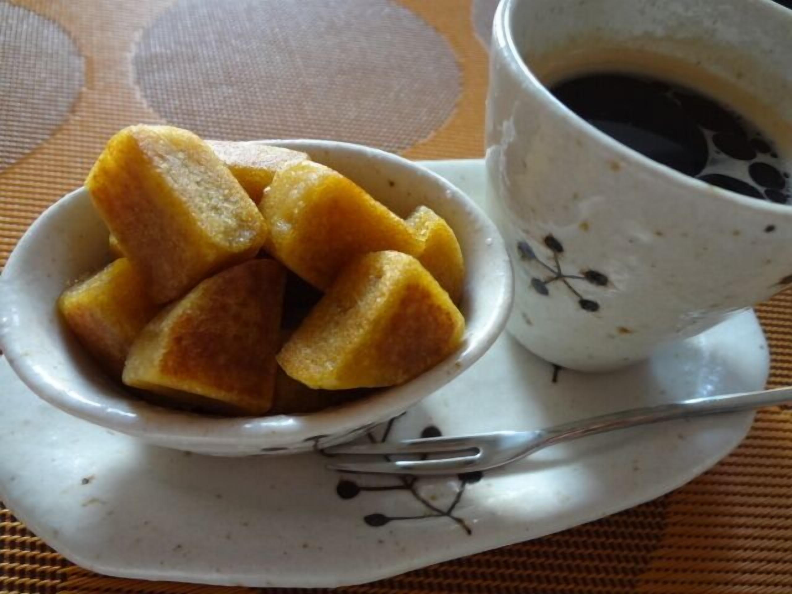 Un tazón con porciones doradas y fritas sobre un plato blanco, junto a una taza blanca con un platito que contiene un líquido oscuro, posiblemente café, sobre una mesa con un posavasos tejido.